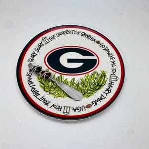 May include: A white ceramic plate with a red and black border. The plate features a green and brown illustration of a football field with the University of Georgia logo in the center. The text "Go Dawgs Sic Em" and "UGA How 'Bout Them Dawgs" is written around the border of the plate.