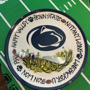 May include: A decorative plate with a Penn State Nittany Lions theme. The plate features the team logo, text, and a decorative knife. The plate has a blue rim and is on a green surface.