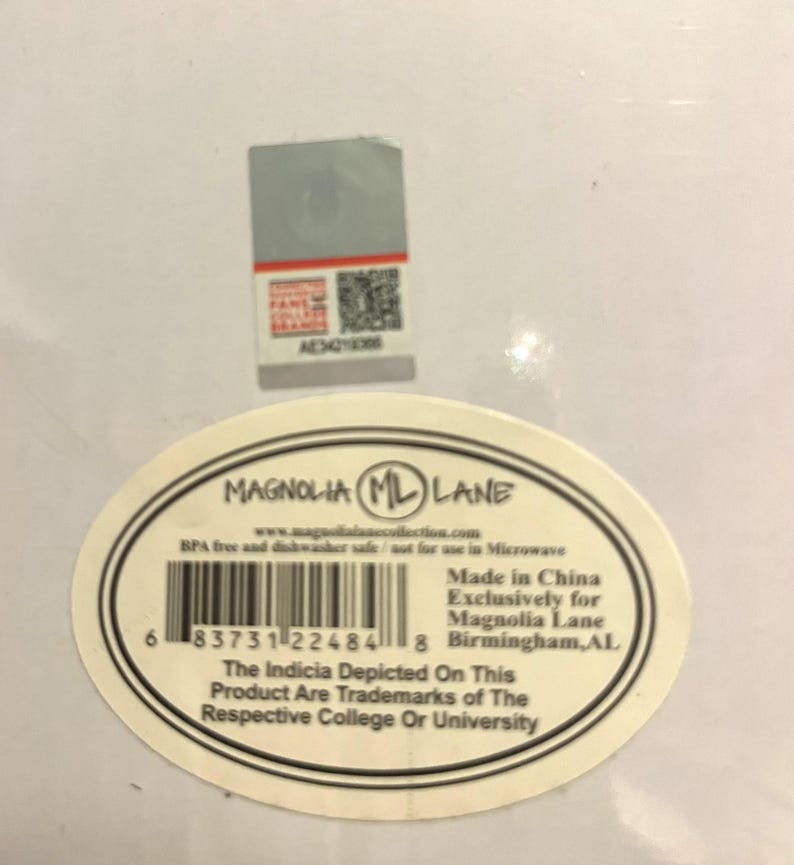 Op de afbeelding: Een wit productlabel met de tekst "MAGNOLIA ML LANE" in een zwarte ovaal. Het label bevat een barcode, websiteadres en tekst die aangeeft dat het product BPA-vrij en vaatwasmachinebestendig is. Een kleinere sticker bevindt zich boven het label.