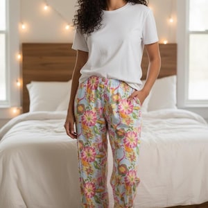 May include: A person wearing a white t-shirt and floral pajama pants. The pajama pants have a light blue background with pink, yellow, and orange floral patterns. The person is standing in a bedroom with a bed and string lights.