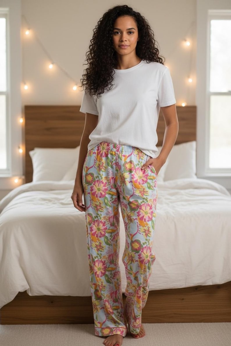 May include: A person wearing a white t-shirt and floral pajama pants. The pajama pants have a light blue background with pink, yellow, and orange floral patterns. The person is standing in a bedroom with a bed and string lights.