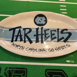 May include: Oval serving platter with a cream-colored base and light blue stripes. The words "TAR HEELS" are prominently displayed in dark blue, with "NORTH CAROLINA: GO HEELS" below. A circular logo is at the top.