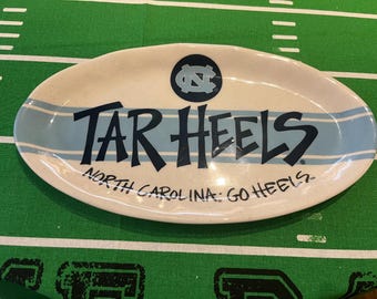 Op de afbeelding: Ovale serveerschaal met een crèmekleurige basis en lichtblauwe strepen. De woorden "TAR HEELS" staan in donkerblauw, met daaronder "NORTH CAROLINA: GO HEELS". Bovenop staat een cirkelvormig logo.