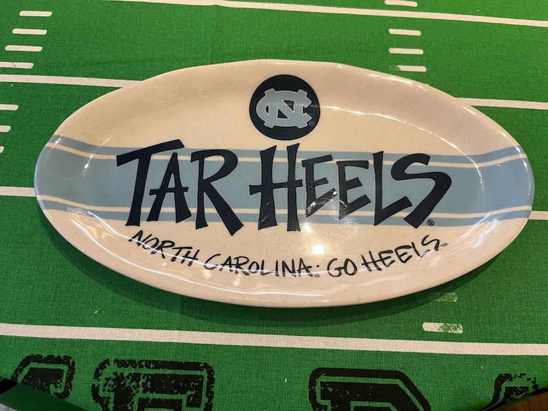 May include: Oval serving platter with a cream-colored base and light blue stripes. The words "TAR HEELS" are prominently displayed in dark blue, with "NORTH CAROLINA: GO HEELS" below. A circular logo is at the top.