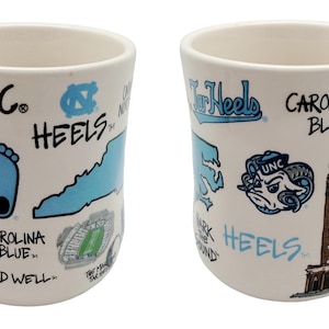 Op de afbeelding: Witte keramische mok met een gebogen handvat, met illustraties en tekst in Carolina blauw en zwart. Het ontwerp bevat de woorden "UNC", "Tar Heels" en "Carolina Blue", samen met een voetafdruk, een ram en een kaart van North Carolina.