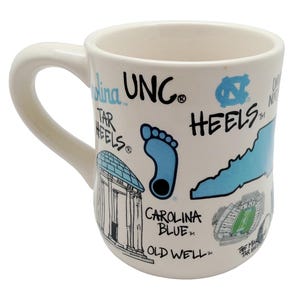 Op de afbeelding: Cr&egrave;mekleurige keramische mok met een gebogen handvat. De mok heeft blauwe en zwarte illustraties en tekst, waaronder een voetafdruk, een staatsomtrek en de woorden "UNC" en "HEELS". De mok heeft ook een illustratie van een gebouw en een stadion.