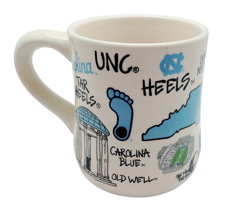 Op de afbeelding: Cr&egrave;mekleurige keramische mok met een gebogen handvat. De mok heeft blauwe en zwarte illustraties en tekst, waaronder een voetafdruk, een staatsomtrek en de woorden "UNC" en "HEELS". De mok heeft ook een illustratie van een gebouw en een stadion.