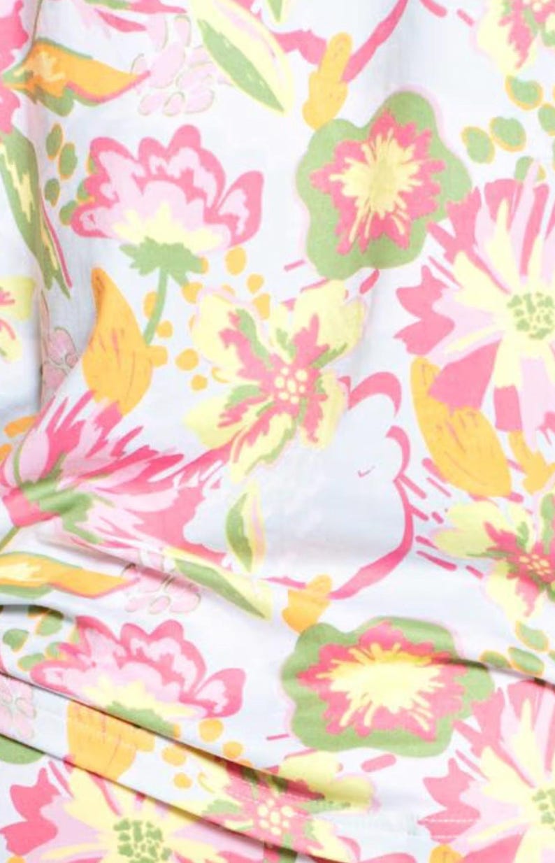 May include: Close-up of a fabric with a floral pattern. The design features large flowers in shades of pink, yellow, and green, set against a light blue background. The fabric has a soft, flowing appearance, suggesting it might be used for clothing or home decor.