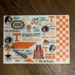 May include: Rectangular cutting board with a Tennessee Volunteers theme. The design includes orange and white checkerboard pattern, team logos, and the words "Tennessee Volunteers", "Rocky Top", and "Go Big Orange".