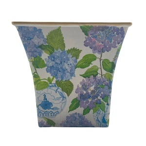 May include: A white ceramic planter with a floral pattern featuring blue and purple hydrangeas and green leaves. The planter has a square shape with slightly flared sides.