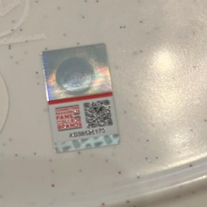 May include: A rectangular, holographic sticker with a red stripe at the top. The sticker features the text "Connecting Fans College Brands" and a QR code. The serial number AD3659173 is printed below the QR code.