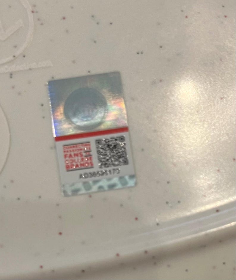 May include: A rectangular, holographic sticker with a red stripe at the top. The sticker features the text "Connecting Fans College Brands" and a QR code. The serial number AD3659173 is printed below the QR code.