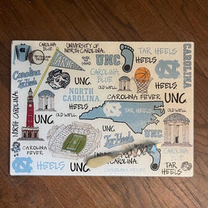 May include: Rectangular cutting board with a UNC Tar Heels theme. The board features illustrations of the North Carolina state outline, a basketball, and the words "Carolina Blue" and "Tar Heels". A corkscrew is also included.