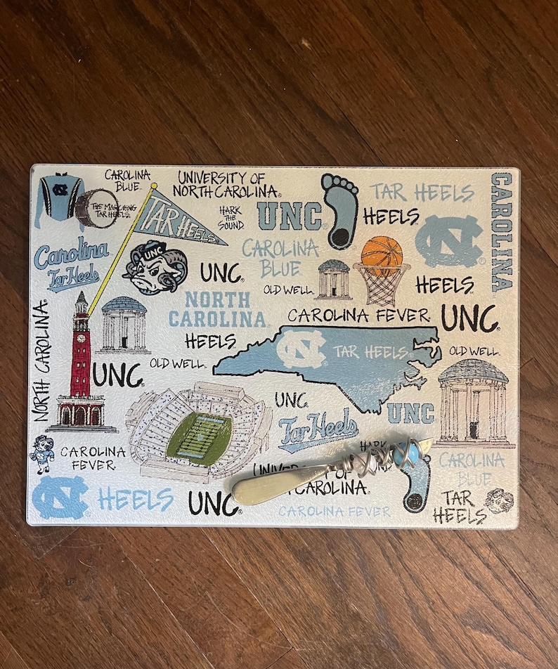 Pu&ograve; includere: Tagliere rettangolare a tema UNC Tar Heels. La tavola presenta illustrazioni del contorno dello stato del North Carolina, un pallone da basket e le parole "Carolina Blue" e "Tar Heels". &Egrave; incluso anche un cavatappi.