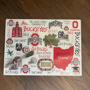 May include: Rectangular glass cutting board featuring a collage of Ohio State University Buckeyes graphics. Includes the Ohio State logo, the state of Ohio in red, and the words "Buckeyes" and "Go Bucks".