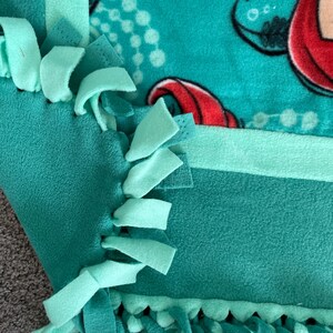 May include: A teal blue and green fleece tie blanket with a mermaid print. The blanket has a fringe border and is made with knotted fabric.
