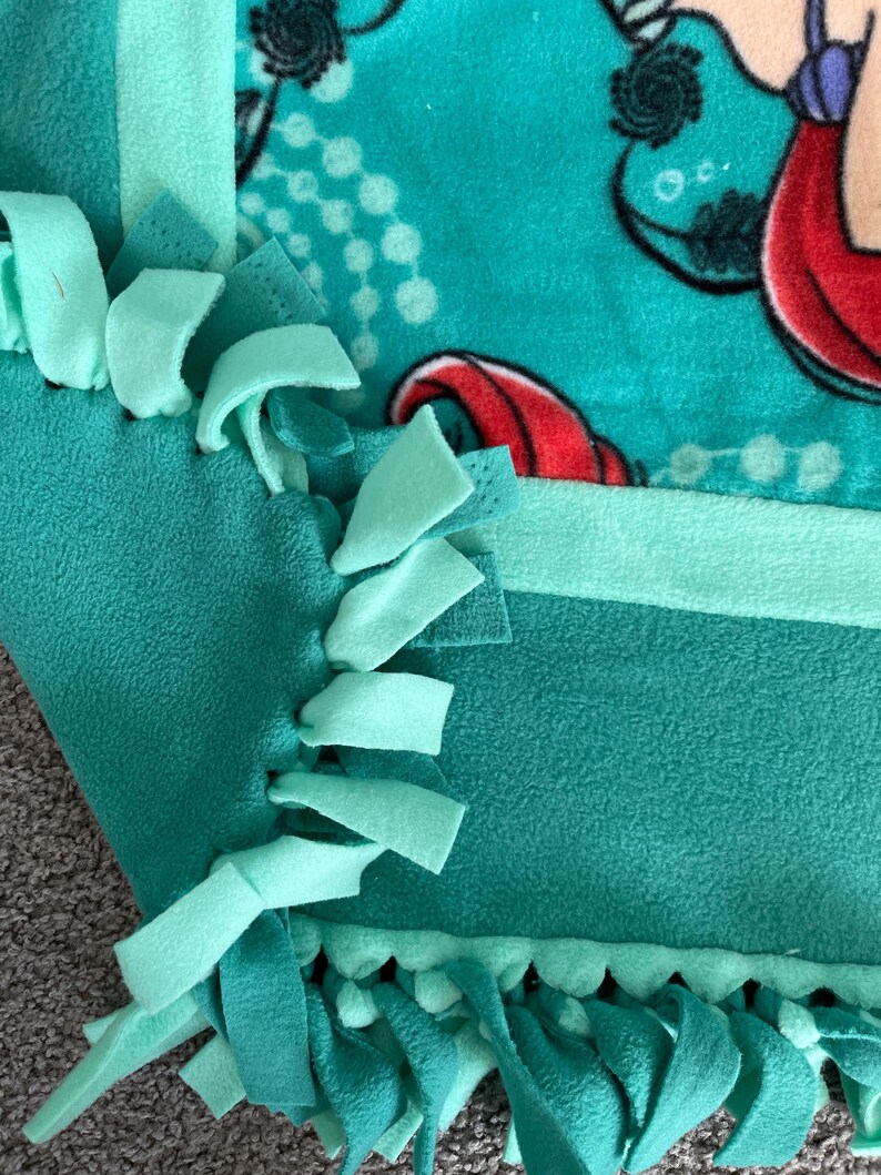 May include: A teal blue and green fleece tie blanket with a mermaid print. The blanket has a fringe border and is made with knotted fabric.
