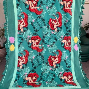 May include: A green fleece tie blanket with a teal border. The blanket features a repeating pattern of Ariel, the Little Mermaid, with the text "Free as the Sea". The blanket has a fringe border and is decorated with yellow and pink felt seashells.