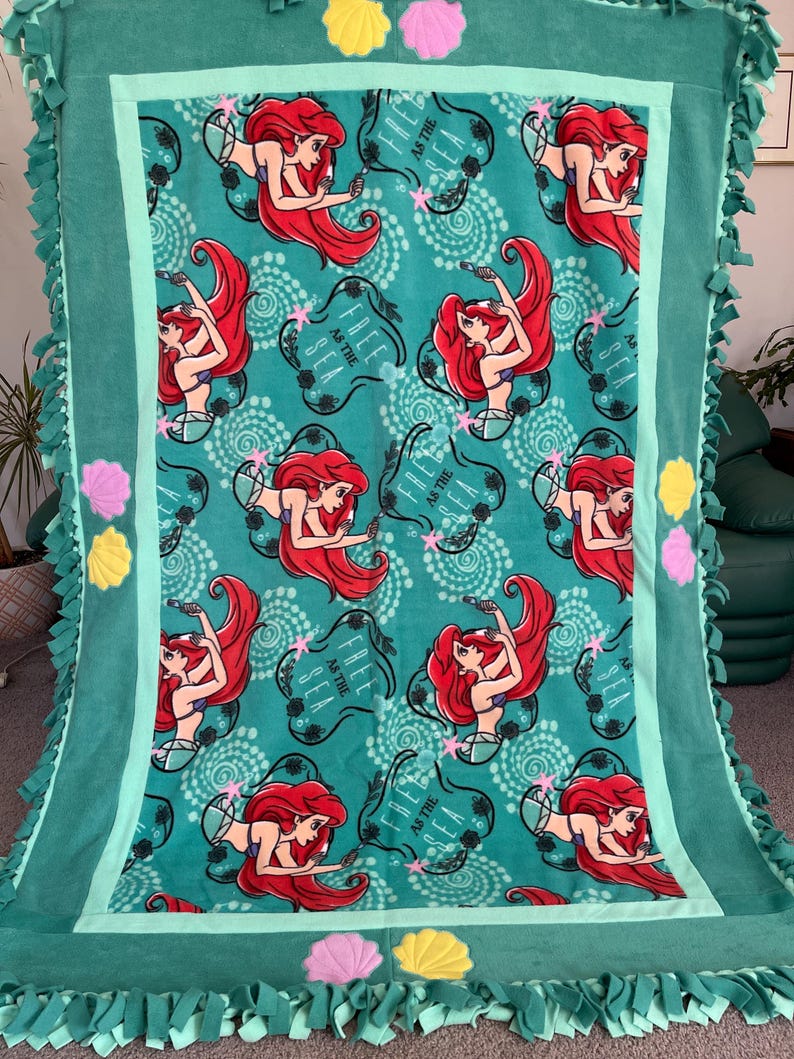 May include: A green fleece tie blanket with a teal border. The blanket features a repeating pattern of Ariel, the Little Mermaid, with the text "Free as the Sea". The blanket has a fringe border and is decorated with yellow and pink felt seashells.