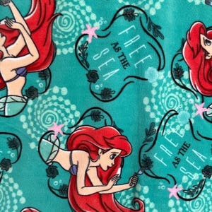 May include: A teal blue fabric with a repeating pattern of Ariel, the Little Mermaid, swimming with a starfish. The text "Free as the Sea" is in a black banner with a floral design.