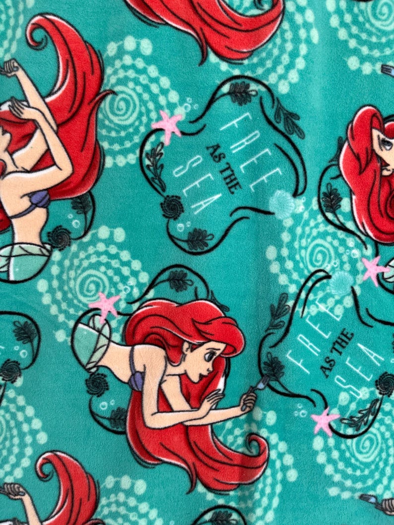 May include: A teal blue fabric with a repeating pattern of Ariel, the Little Mermaid, swimming with a starfish. The text "Free as the Sea" is in a black banner with a floral design.