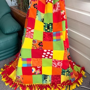 May include: A colorful patchwork fleece blanket with a variety of patterns and colors, including red, orange, yellow, and green squares. The blanket has a fringed edge with red and yellow accents. The blanket is draped over a chair.