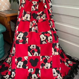 May include: Red fleece blanket with a patchwork design featuring Mickey Mouse faces. The blanket has black fringe around the edges.