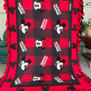 May include: Red and black fleece tie blanket with a checkered pattern and Mickey and Minnie Mouse images. The blanket has black fringe around the edges.