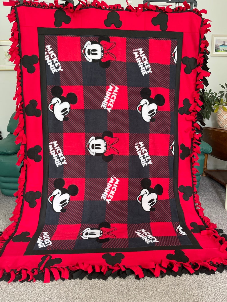 May include: Red and black fleece tie blanket with a checkered pattern and Mickey and Minnie Mouse images. The blanket has black fringe around the edges.