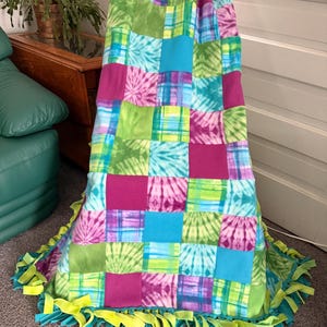 May include: A colorful patchwork fleece blanket with a tie-dye pattern in shades of pink, blue, and green. The blanket has a fringed edge in teal and neon yellow. The blanket is draped over a chair.