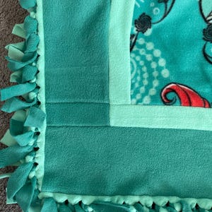 May include: A turquoise blue fleece blanket with a green border and a knotted fringe. The blanket has a light green border and a darker green border.