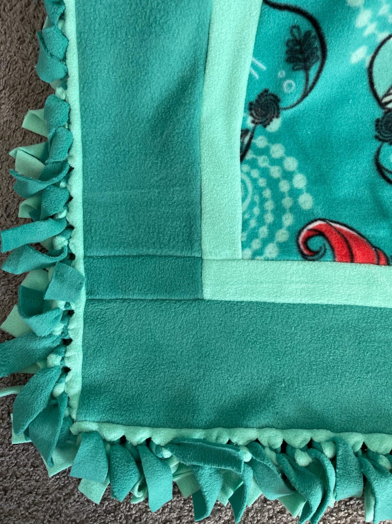 May include: A turquoise blue fleece blanket with a green border and a knotted fringe. The blanket has a light green border and a darker green border.