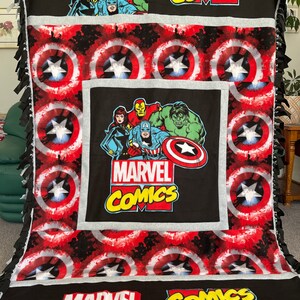 May include: A black and red fleece blanket with a Marvel Comics theme. The blanket features a repeating pattern of Captain America's shield, and a large image of Marvel characters including Captain America, Iron Man, Hulk, and Black Widow. The blanket also has the words "Marvel Comics" printed on it.
