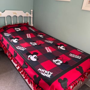 May include: A red and black fleece blanket with a Mickey Mouse and Minnie Mouse pattern. The blanket has a knotted fringe around the edges.