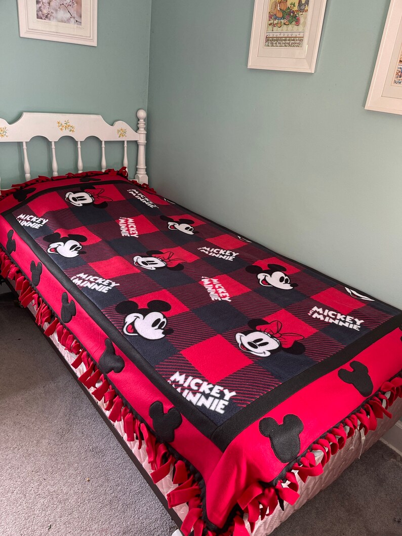 May include: A red and black fleece blanket with a Mickey Mouse and Minnie Mouse pattern. The blanket has a knotted fringe around the edges.