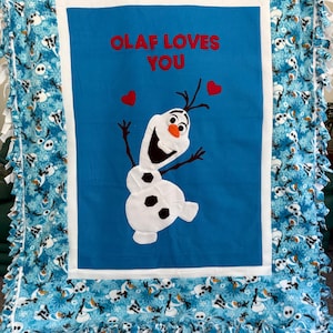 May include: A blue and white fleece blanket featuring Olaf from Frozen. The center has a blue square with Olaf and the text "OLAF LOVES YOU" in red. The blanket has a white border and a fringe edge.