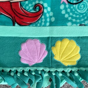 May include: A green fleece tie blanket with a pink and yellow seashell applique. The blanket has fringe on the bottom edge.