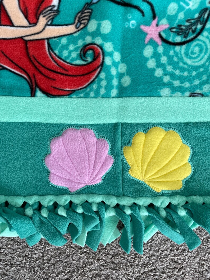 May include: A green fleece tie blanket with a pink and yellow seashell applique. The blanket has fringe on the bottom edge.