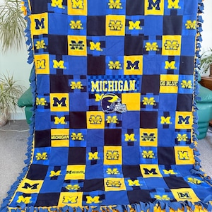 May include: A blue, yellow, and black fleece blanket featuring the word "MICHIGAN" and the letter "M" repeated throughout. The blanket has a grid pattern with a football helmet graphic and the words "GO BLUE". The edges are fringed.