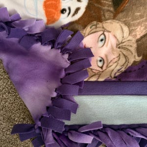 以下が含まれることがあります： 映画「アナと雪の女王」のキャラクターが描かれたフリースブランケット。ブランケットは、紫色のグラデーションの縁取りと結び目のフリンジが付いています。キャラクターのオラフとエルサがブランケットに描かれています。