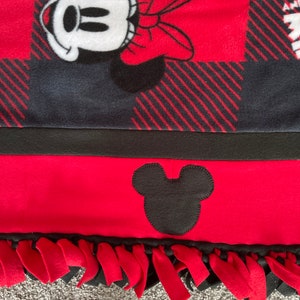 May include: Red and black fleece blanket with a Mickey Mouse design. The blanket has a black and red plaid pattern and a black Mickey Mouse head on the bottom. The blanket has red fringe around the edges.