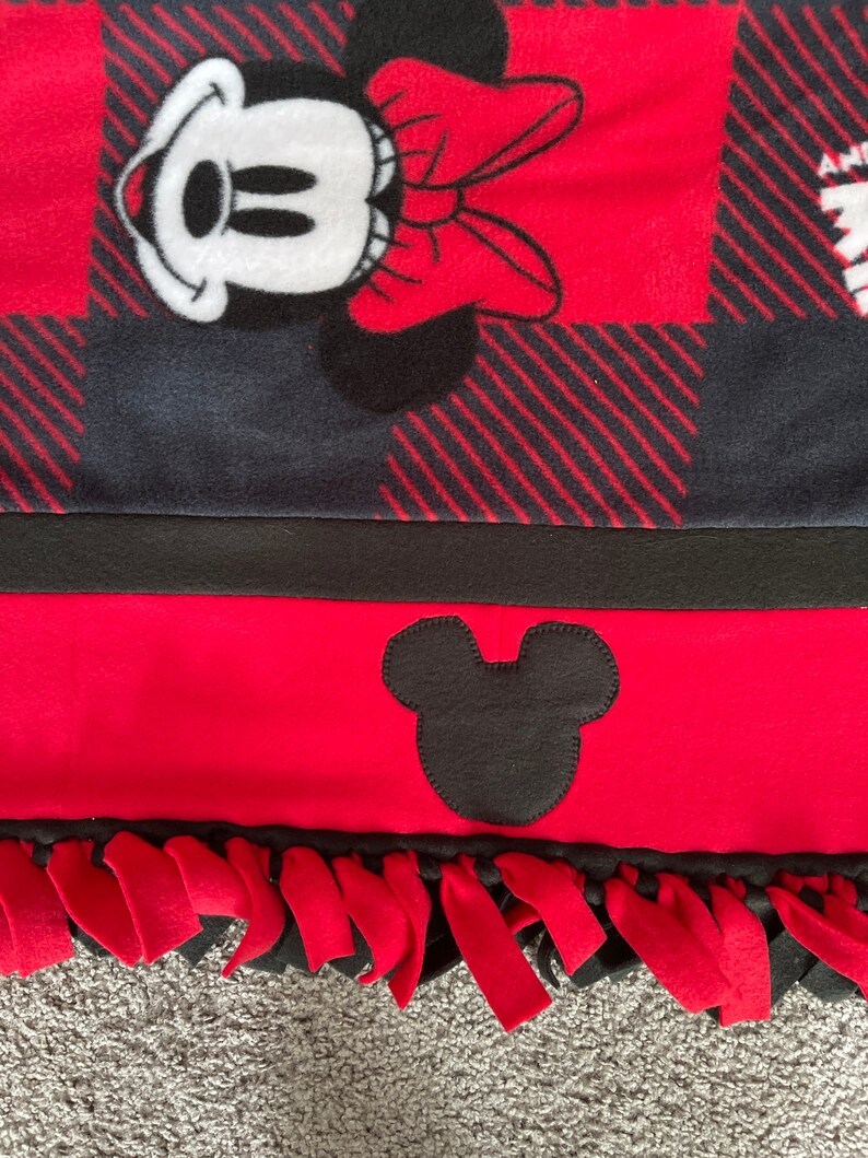 May include: Red and black fleece blanket with a Mickey Mouse design. The blanket has a black and red plaid pattern and a black Mickey Mouse head on the bottom. The blanket has red fringe around the edges.