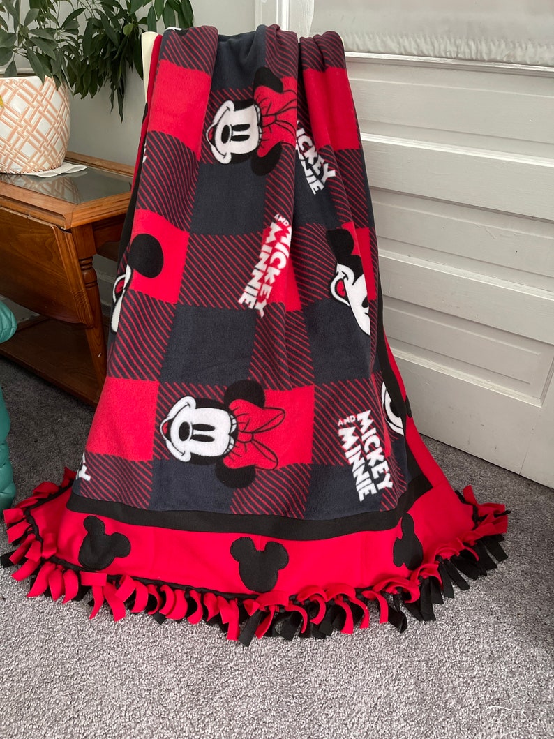May include: Red and black fleece blanket with a checkered pattern and Mickey and Minnie Mouse images. The blanket has a knotted fringe border.