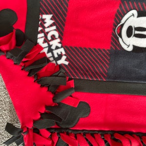 May include: A red and black fleece blanket with Mickey Mouse ears and the word "Mickey" on it. The blanket has black and red fringe around the edges.