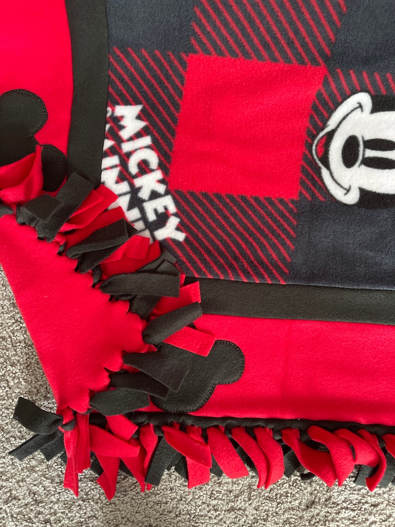 May include: A red and black fleece blanket with Mickey Mouse ears and the word "Mickey" on it. The blanket has black and red fringe around the edges.