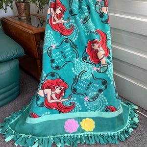 May include: A teal blue fleece blanket with a repeating pattern of Ariel, the Little Mermaid, with the text "Free as the Sea". The blanket has a teal blue fringe and two embroidered seashells in pink and yellow.