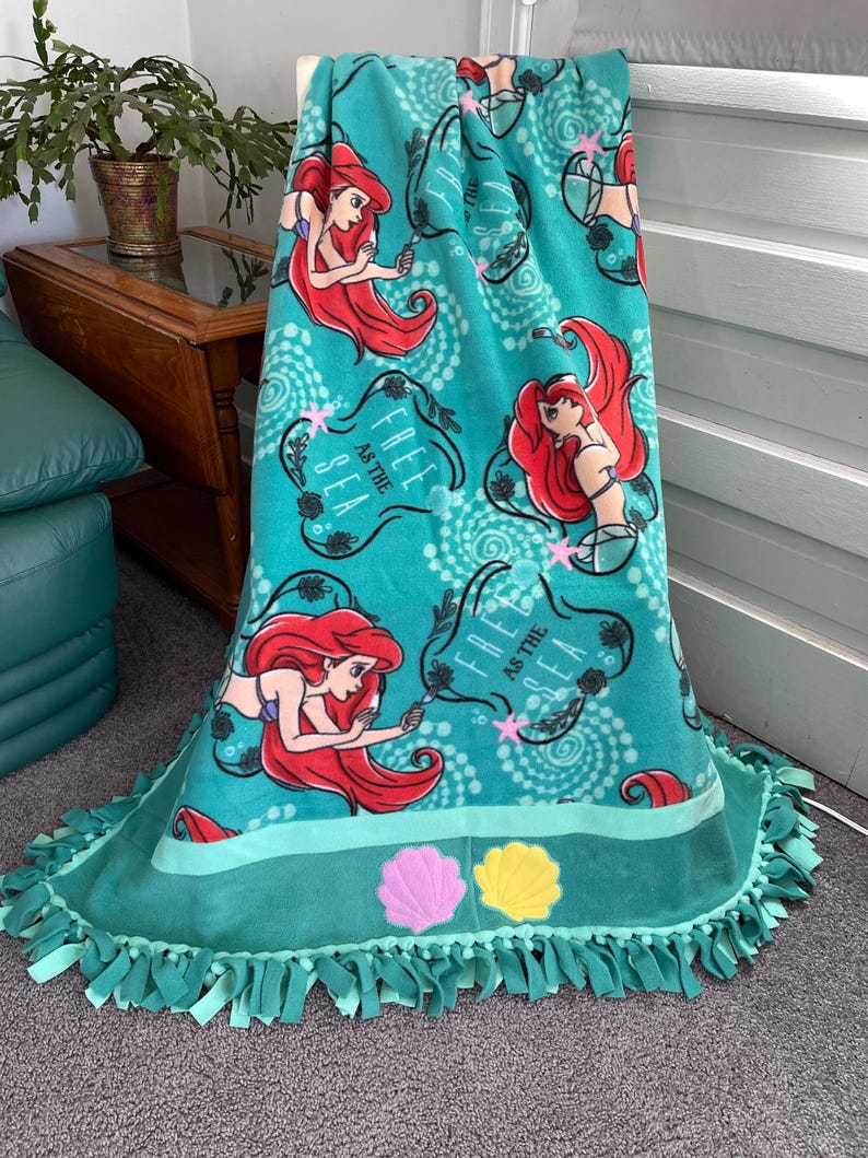 May include: A teal blue fleece blanket with a repeating pattern of Ariel, the Little Mermaid, with the text "Free as the Sea". The blanket has a teal blue fringe and two embroidered seashells in pink and yellow.