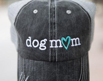 May include: A black and gray trucker hat with the words "dog mom" embroidered in white, with a teal heart replacing the "o". The hat has a distressed look and a gray mesh back.