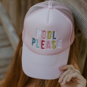 May include: A light pink trucker hat with a mesh back and a curved brim. The hat features the embroidered words "POOL PLEASE" in colorful letters. A fun accessory for the pool or beach.