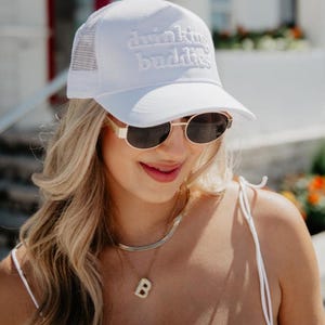 May include: A white trucker hat with the text "drinking buddies" embroidered on the front. The hat has a mesh back and a curved brim. The person is wearing gold-rimmed sunglasses and a gold necklace with a "B" pendant.